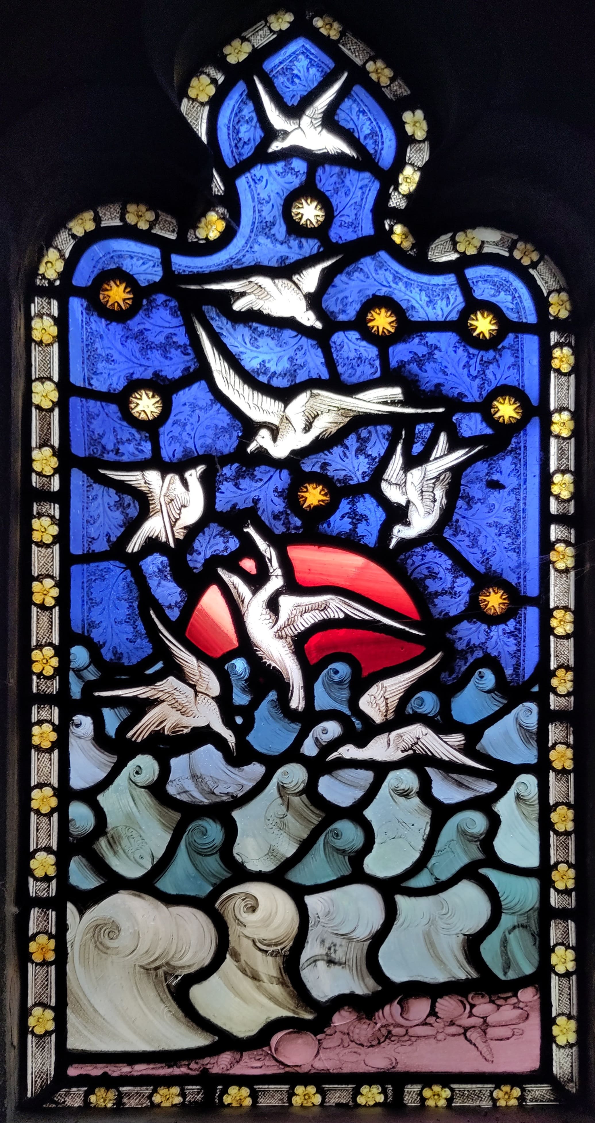 An image of a stained glass window. It depicts a red sun setting over the sea, with stars in the sky. A flock of seabirds are flying in front